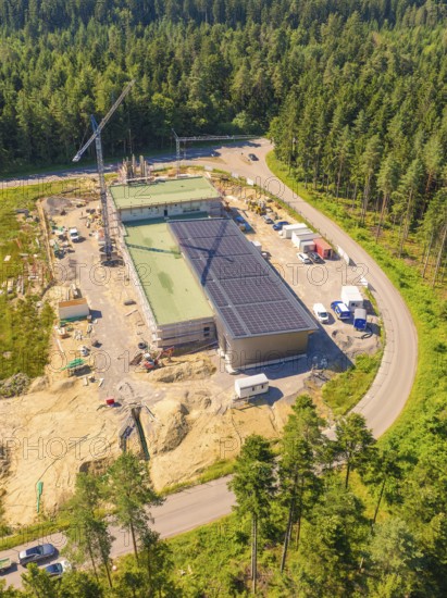 Building with solar roof in a construction site surrounded by forest, crane nearby, various vehicles and containers visible, new fire brigade building, Oberhaugstett, Calw district, Germany