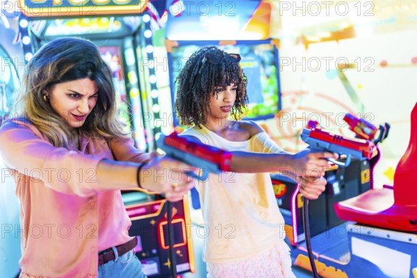 Two young women focusing intently while enjoying a thrilling shooting game in a vibrant amusement arcade filled with colorful lights