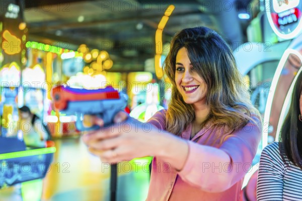 Cheerful young woman holding a toy gun, playing at an arcade game machine, enjoying her free time with friends