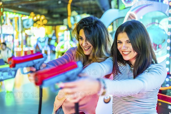 Two young women enjoying leisure time, playfully engaging with toy guns in a vibrant amusement arcade filled with colorful lights and laughter