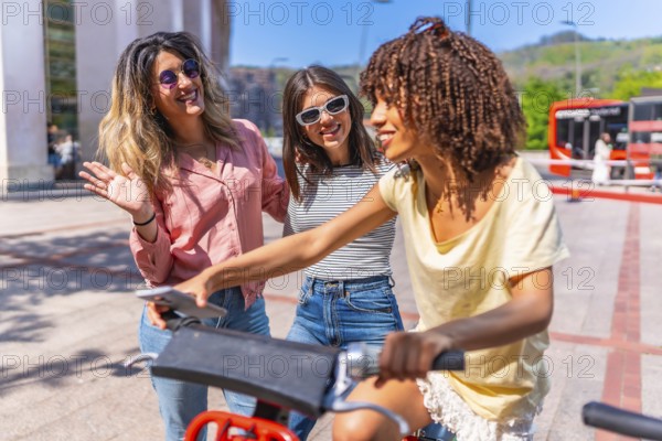 Three cheerful young women using smartphone and riding a sharing bike in a city center during summer holidays