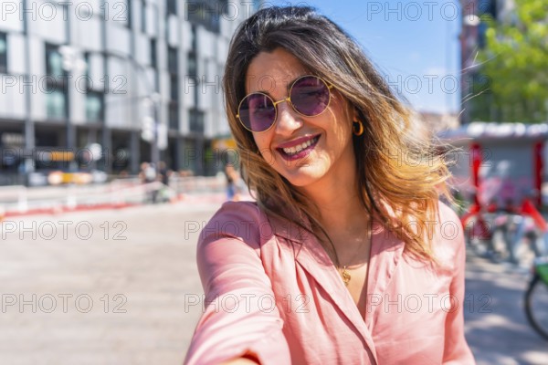 Smiling young woman wearing trendy sunglasses, capturing a joyful selfie in a vibrant city center on a sunny summer day