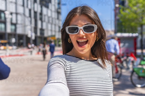 Happy woman wearing trendy sunglasses, joyfully taking a selfie in the bustling city center on a bright summer day
