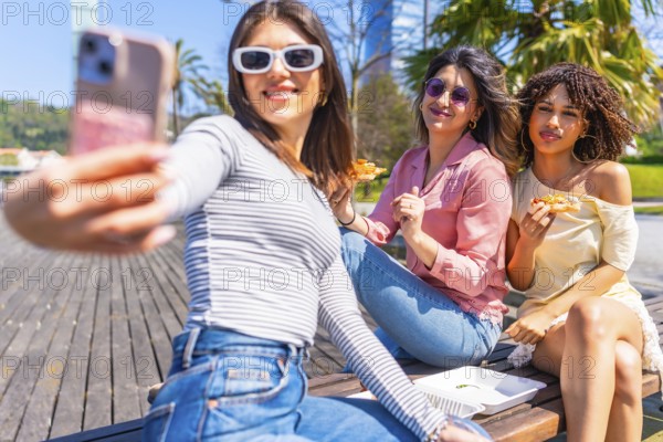 Group of female friends taking a selfie while enjoying pizza on a bench in a city park during a sunny summer day
