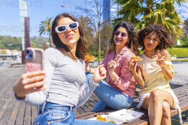 Three happy women taking a selfie while eating pizza on a bench in the city during a sunny day