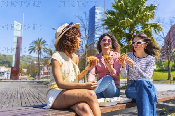 Three women savoring pizza slices while enjoying a sunny day on a park bench, sharing laughter and creating lasting memories in the city