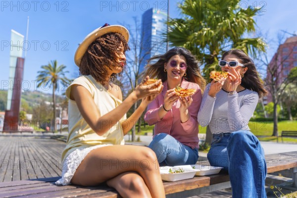 Three young women savoring delicious pizza while sitting on a bench in a vibrant city park, enjoying a sunny summer day together