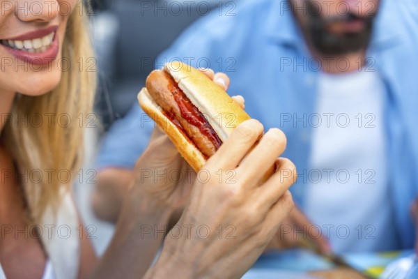Woman enjoying a hot dog with ketchup, sitting at a picnic table, with a blurred man in the background