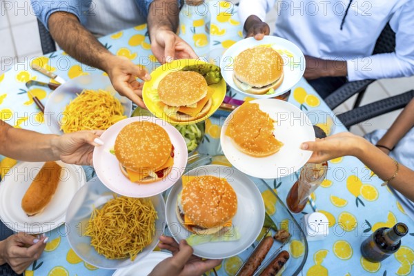 Hands holding plates with hamburgers and fries during a barbecue with friends, sharing food on a table with a lemon patterned tablecloth