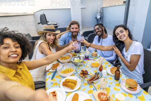 Cheerful multi ethnic friends enjoying rooftop barbecue party, taking selfie and toasting with drinks