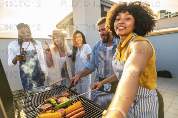 Cheerful friends grilling delicious meat and fresh vegetables while capturing selfies at a vibrant sunset party on the rooftop