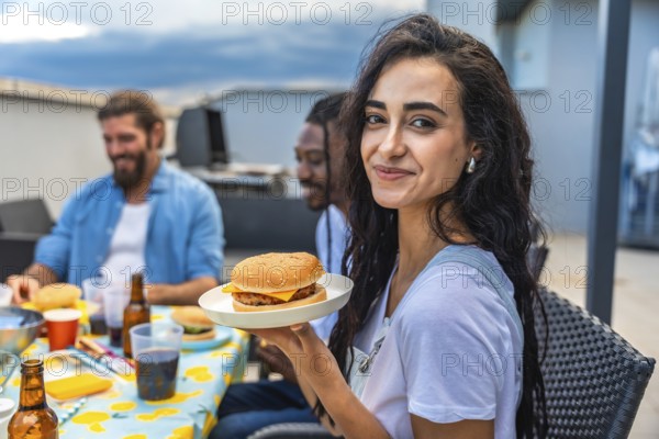 Young woman savoring a delicious burger while celebrating with friends at a lively rooftop barbecue party, enjoying the summer vibes