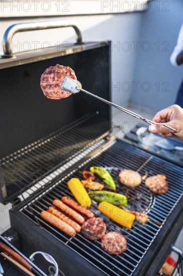 Hand using tongs to grill hamburgers, sausages, corn and peppers on a barbecue grill during a summer party