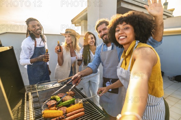 Cheerful multi ethnic friends are grilling meat and vegetables, drinking beer and taking a selfie on a rooftop at sunset