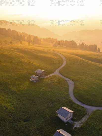 Mountain huts on gentle hills with scattered groups of trees in the light of the sunset, Alpe di Siusi, Dolomites, South Tyrol, Italy