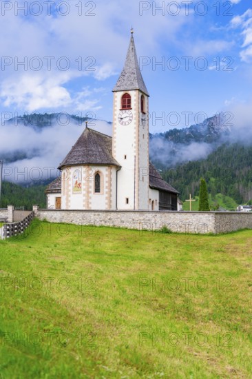 Church with clock tower in a green alpine landscape under a cloudy sky, Dolomites, South Tyrol, Italy