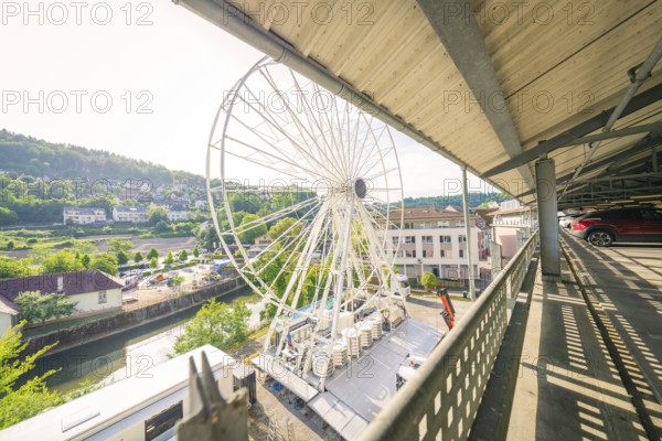 A Ferris wheel stands next to a river under the roof of a multi-storey car park, surrounded by buildings and green trees, Ferris wheel construction, Calw, Black Forest, Germany