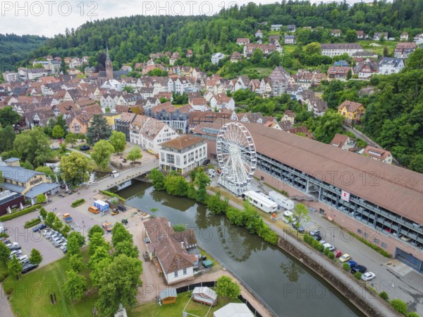 A cityscape with a Ferris wheel on a river, surrounded by a dense ensemble of buildings and hills, Ferris wheel structure, Calw, Black Forest, Germany