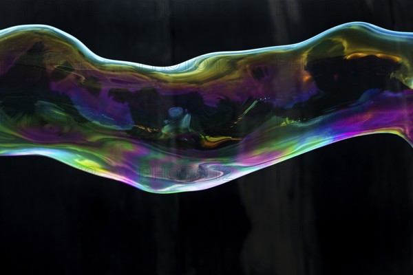 Large, iridescent soap bubble tube in the colours of the rainbow against a black background, Recklinghausen, Germany