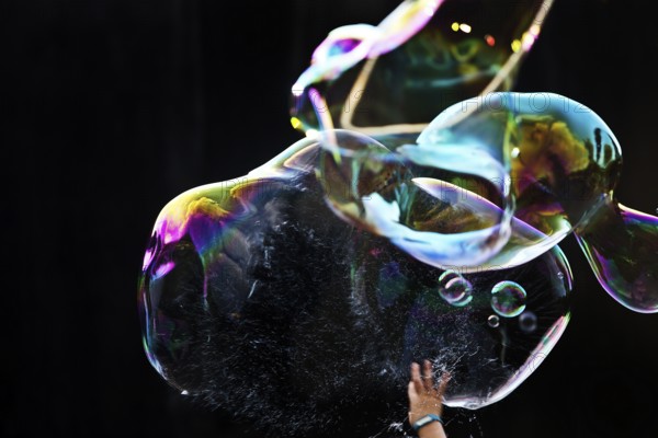 A hand bursts a large iridescent soap bubble against a black background, Recklinghausen, Germany