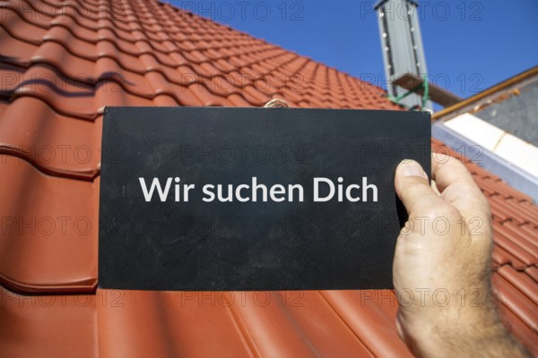 Employee search roofer: Man holding a sign (with added text) WE ARE LOOKING FOR YOU in front of a new tiled roof
