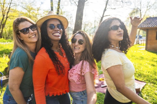 Four diverse women wearing stylish sunglasses and sharing joyful smiles while relaxing together in a sunny park on a beautiful day