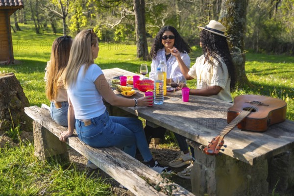Four young women are having a picnic on a wooden table, with wine, fruit, and a guitar, enjoying the sunny day in a park