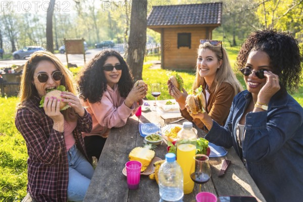 Four young women enjoying a picnic lunch outdoors, eating hamburgers and sharing good times together