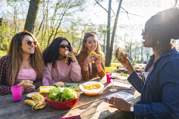 Group of young women enjoying a sunny picnic at a park, happily sharing delicious sandwiches and laughter around a picnic table