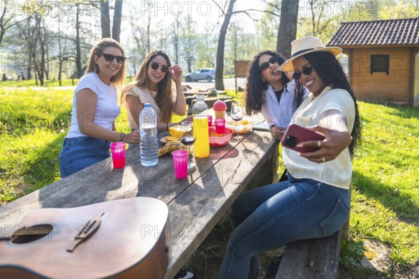 Four young women taking a selfie with smartphone at a picnic table in a park during a sunny day, with guitar, fruit and drinks