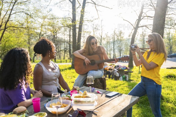 Four female friends enjoying a picnic in a park, with one playing guitar, another taking photos, and the others relaxing at the table