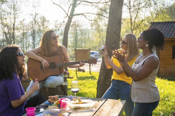 Four friends are having a picnic outdoors, with one playing guitar, one taking photos, and the others enjoying the music and food
