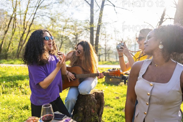 Four young women enjoying a picnic in a park, singing, playing guitar, taking pictures, and having fun
