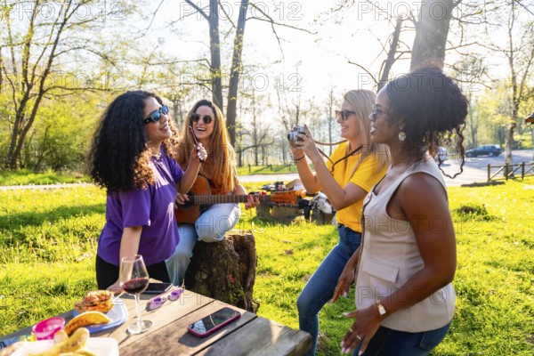 Four cheerful female friends are having a picnic in a sunny park, playing guitar, taking photos, and enjoying each other's company