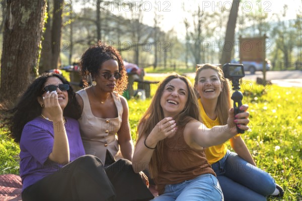 Four multi ethnic female friends are sitting on grass in a park, taking a selfie with an action camera mounted on a gimbal