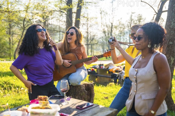 Four female friends enjoying a picnic in a park, singing, playing guitar, taking photos, and having fun