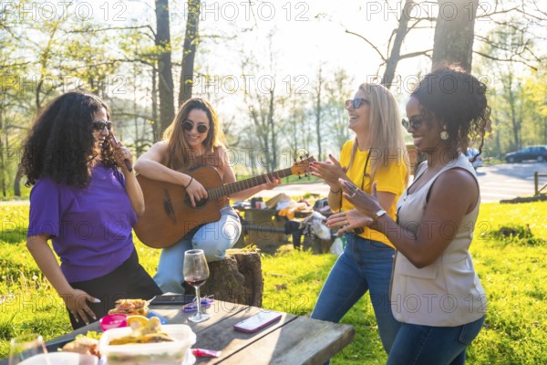 Four diverse female friends are enjoying a picnic in a park, singing, playing guitar, and clapping their hands while celebrating friendship