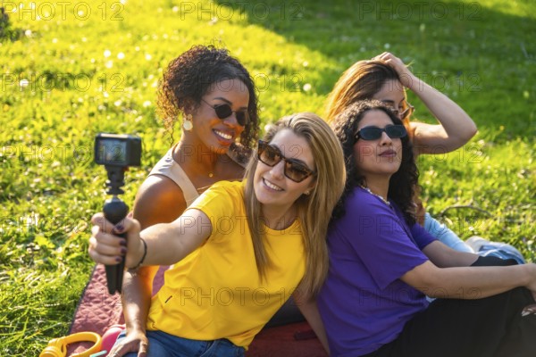 Four cheerful female friends are recording a video blog in the park using an action camera, enjoying their time together on a sunny day