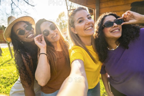 Four friends are taking a selfie outdoors, wearing sunglasses and smiling, enjoying a sunny day and having fun together