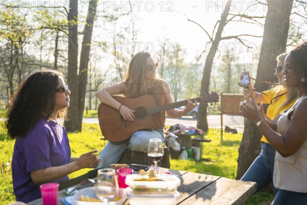 Group of friends enjoys a picnic in a park, with one playing guitar while another takes photos