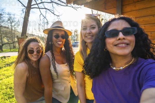 Four diverse women wearing sunglasses and smiling while taking a selfie together in a sunny park