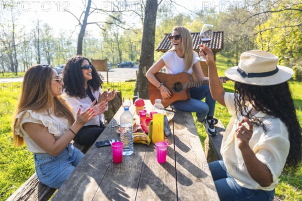Four diverse women are having a picnic in a park, playing guitar, clapping, drinking wine, and enjoying the sunny weather
