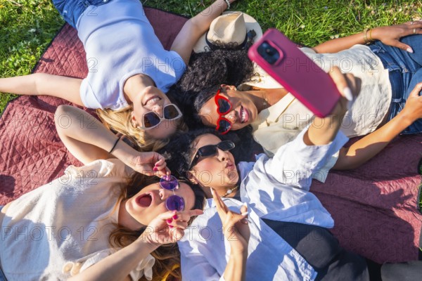 Four cheerful female friends are lying on a blanket in a park, taking a selfie with a smartphone and making funny faces