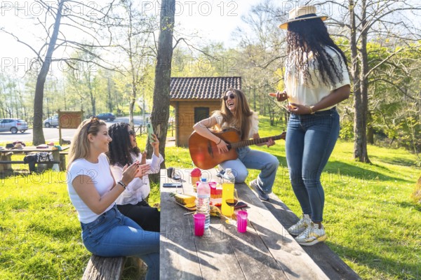 Four friends are having a picnic in a park, with one playing guitar, another dancing, and two others clapping and recording