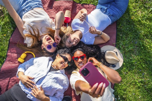 Four female friends are relaxing on a blanket in a park, taking a selfie with a smartphone on a sunny summer day