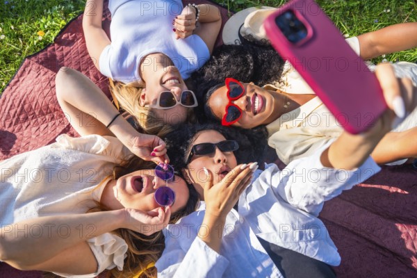 Four cheerful young women wearing sunglasses are lying on a blanket in a park, taking a selfie with a smartphone