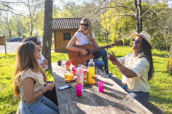 Happy friends are having a picnic in a park, enjoying music played on an acoustic guitar, sharing food and drinks on a sunny day