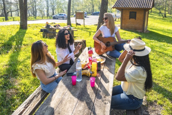 Four female friends enjoying a sunny picnic in nature, playing guitar, singing joyfully, and sharing delicious food together