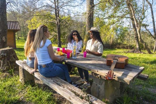 Group of female friends enjoying picnic lunch and drinks at wooden table in sunny forest park with acoustic guitar
