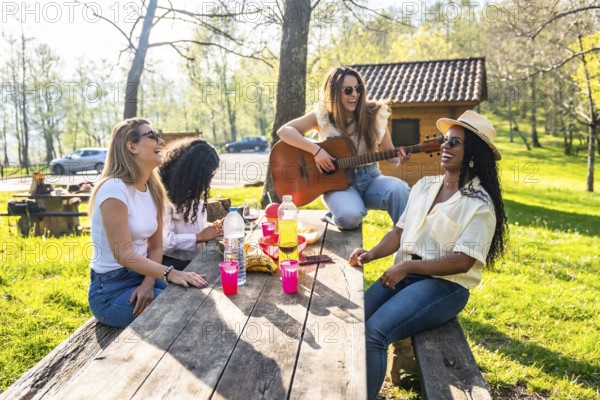 Four female friends are sitting at a wooden table, playing guitar and enjoying a picnic outdoors, with drinks and snacks